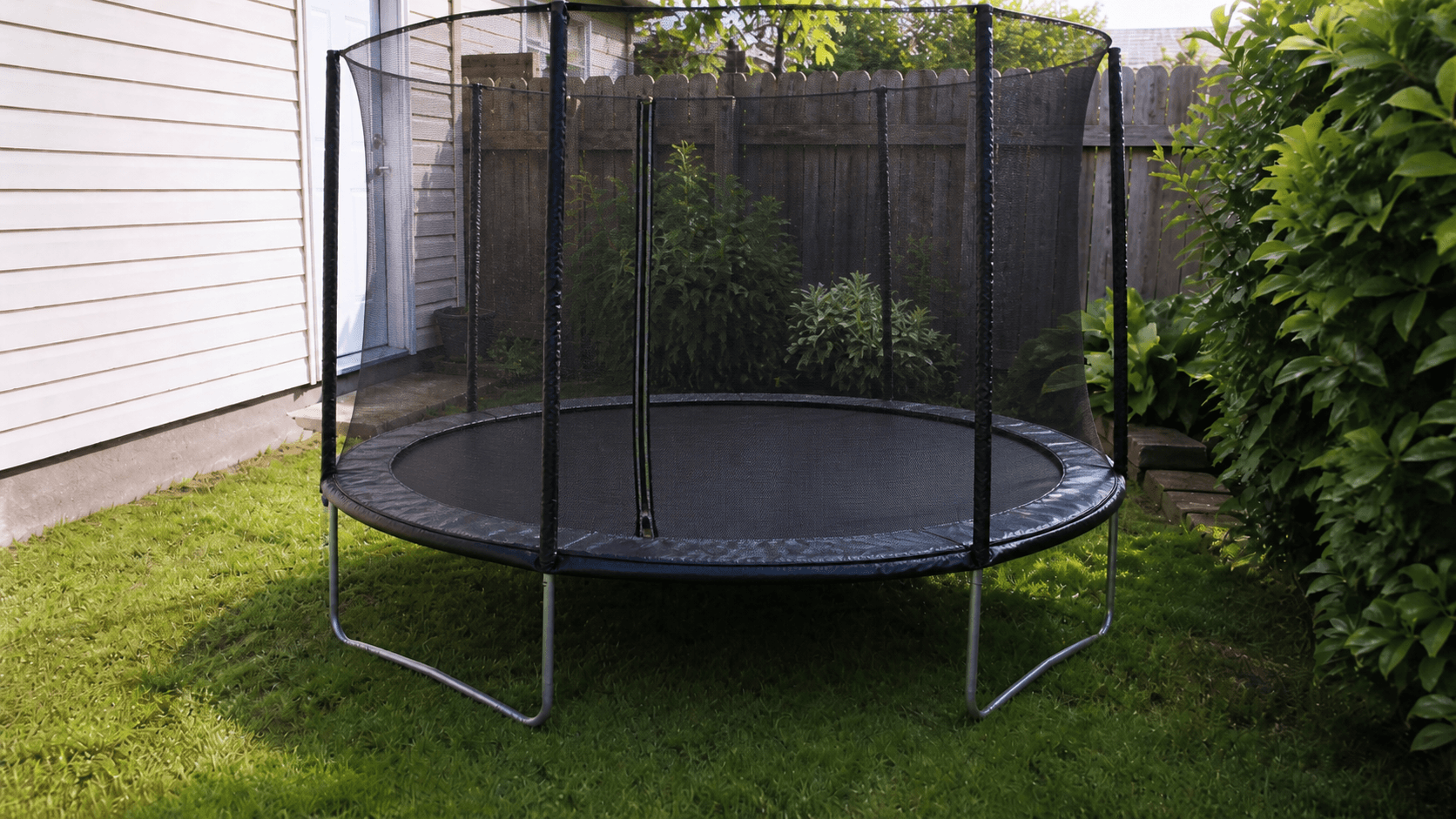 A small enclosed trampoline fitted into a narrow backyard beside a house.